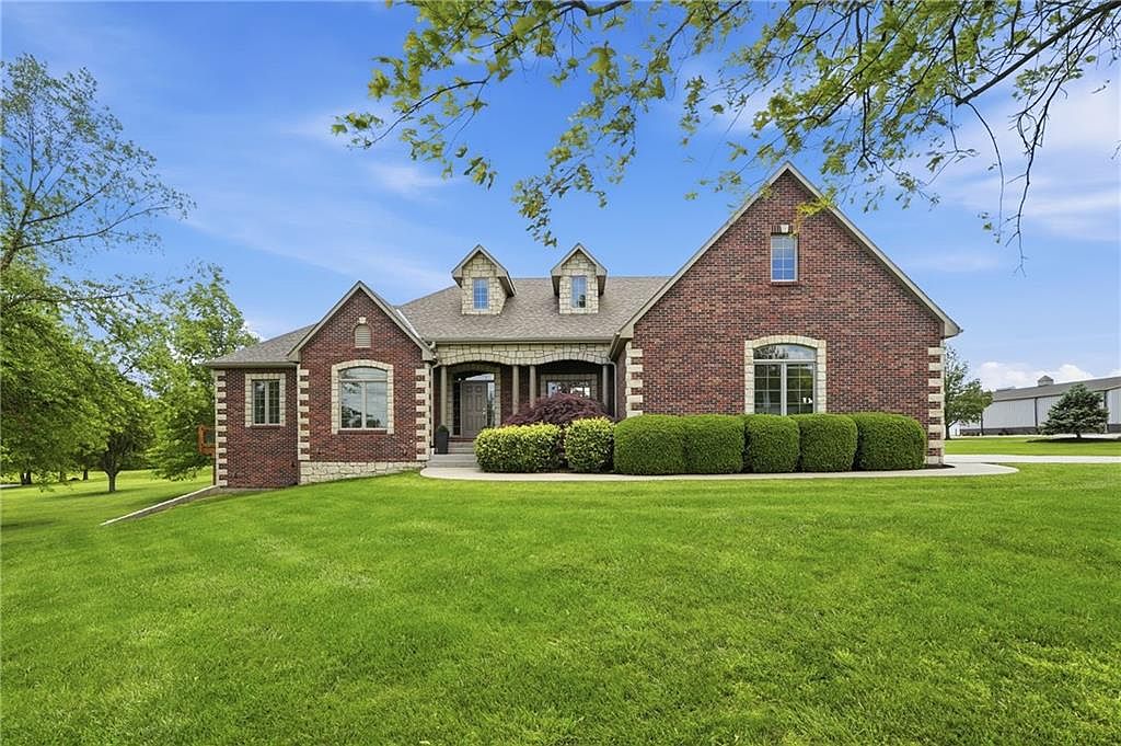 Front elevation of 26619 K68 Hwy, an all-brick one-level estate home with cedar-shake gable dormers, stone-trimmed entry, and manicured hedges on a generous lawn outside Paola, Kansas